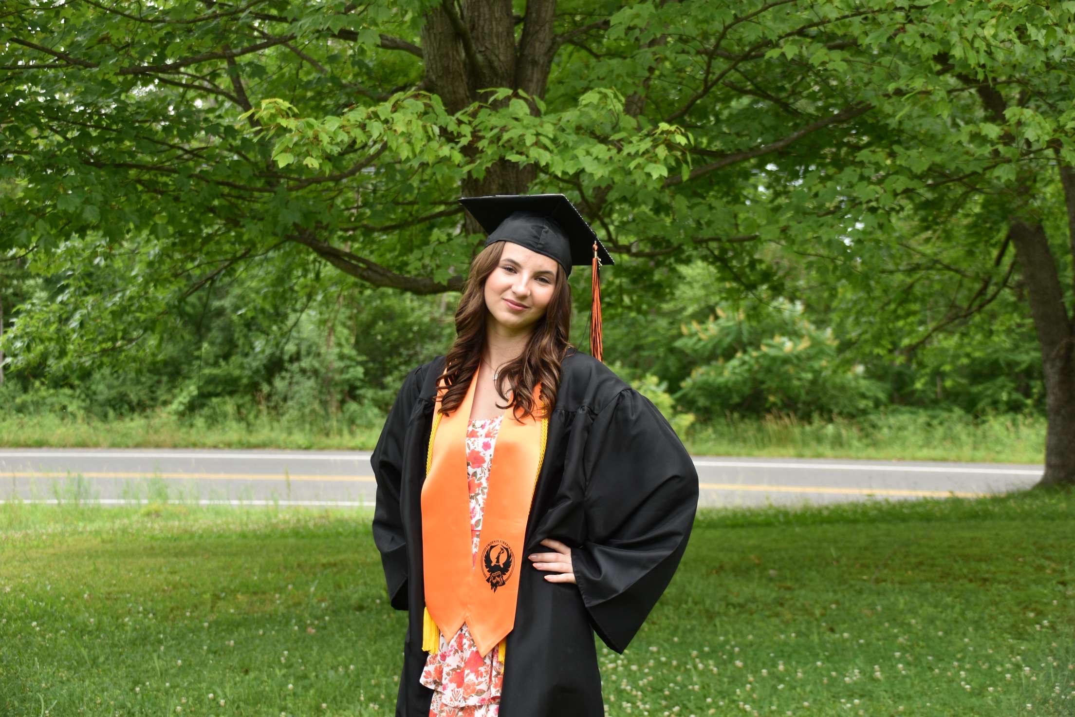 Young woman in a graduation cap and gown and orange sash standing in front of a scenic background