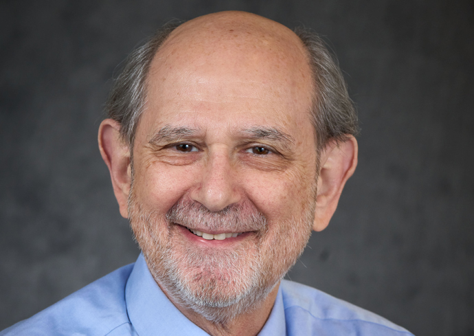 A portrait of a person who has gray hair and is wearing a light blue collared shirt. The background is dark and plain.