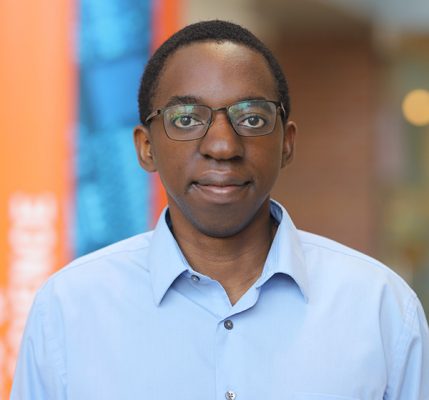A young persom with short hair and black-rimmed glasses looks directly at the camera with a slight smile. They are wearing a light blue button-down dress shirt. The background shows a modern, colorful interior space with blurred orange and blue architectural elements, suggesting an office or contemporary building environment.