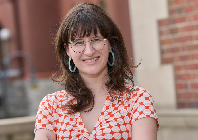 A person with long brown hair wearing a short-sleeved shirt with an orange and white circular pattern and large hoop earrings, standing outdoors in front of a blurred red brick building.