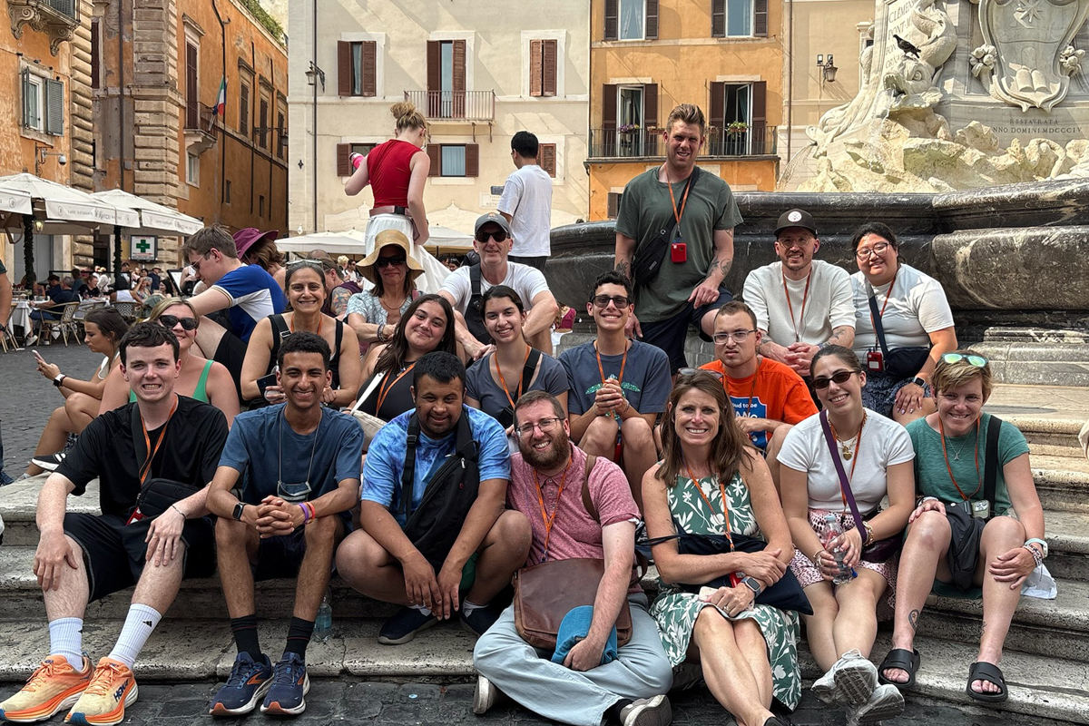 A large group of young adults, appearing to be college students or study abroad participants, poses together on the stone steps of an ornate fountain in what appears to be an Italian piazza. The group of approximately 15-20 people is arranged in multiple rows, all wearing colorful summer clothing and many sporting orange lanyards, suggesting they're part of an organized program or tour. Behind them stands an elaborate baroque-style fountain with carved stone figures and decorative elements. The scene is set in a historic European square with characteristic orange and cream-colored buildings featuring traditional shuttered windows and balconies. Other tourists can be seen in the background, and the cobblestone plaza and architecture suggest this is likely Rome or another major Italian city.