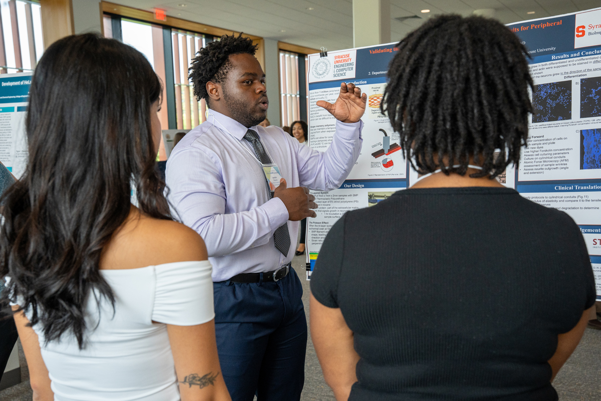 A person in a white dress shirt and tie presents research at an academic poster session, gesturing with his hand while explaining his work to two attendees.