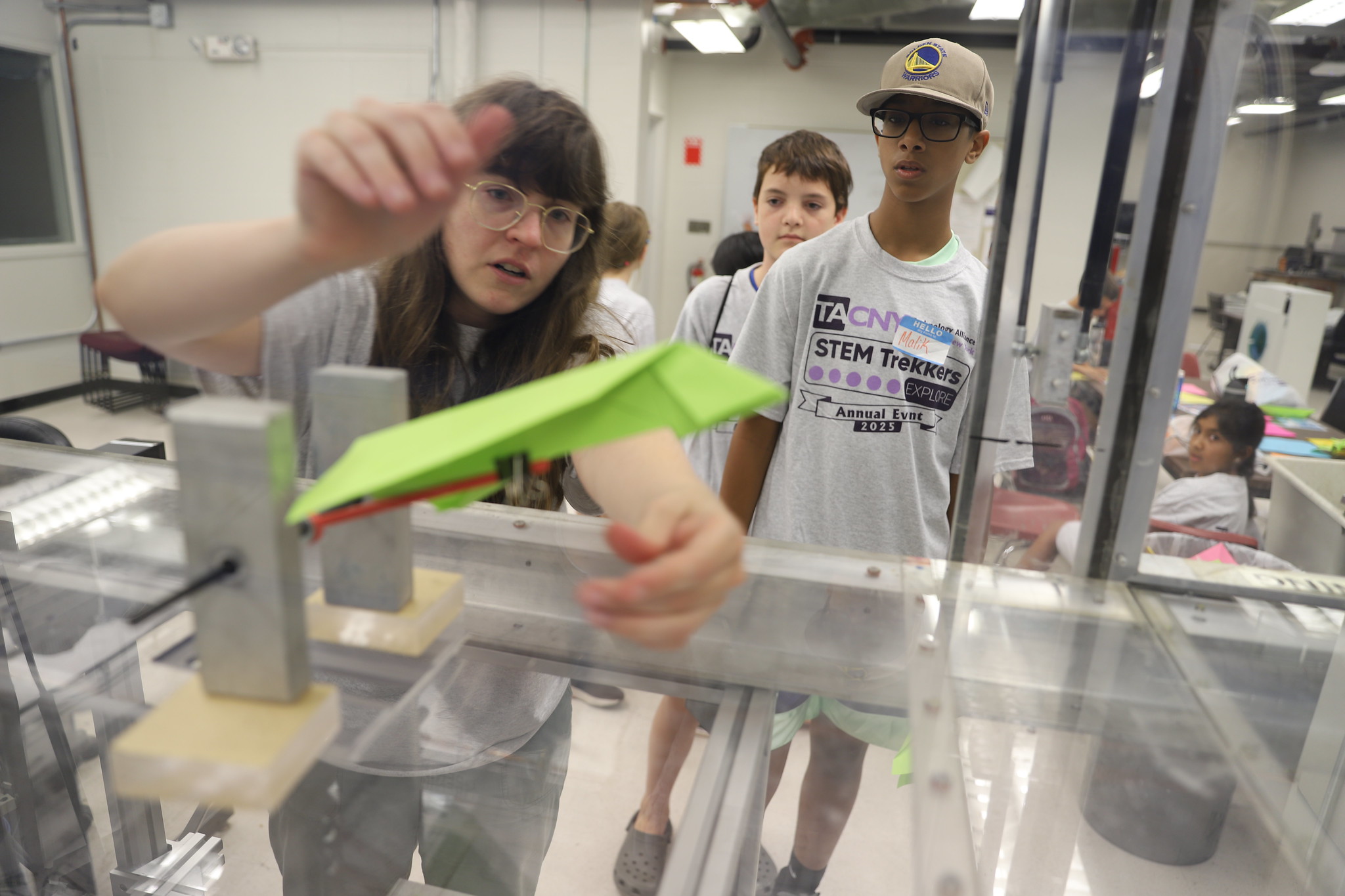 A person with glasses and long brown hair reaches toward a bright green paper airplane or similar object inside what appears to be a clear plastic testing chamber or wind tunnel. They are wearing a gray shirt and is focused on the experiment. Behind her, two other students observe - one wearing a Golden State Warriors cap and a "STEM Trekkers" t-shirt, and another student in the background. The setting appears to be a science laboratory or classroom with white walls, fluorescent lighting, and various equipment visible.