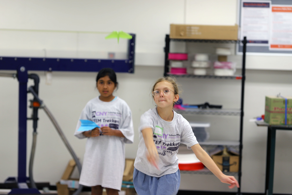Two young students participate in a paper airplane activity in a classroom setting. In the foreground, a student with glasses and blonde hair pulled back extends their arm, appearing to have just thrown or be about to throw a paper airplane. They are wearing a white "STEM Trekkers" t-shirt and a blue skirt. Behind them, another student with dark hair also wears a white "STEM Trekkers" shirt and holds a blue paper airplane. A bright green paper airplane is visible flying through the air in the upper left portion of the image. The classroom has white walls, storage shelves with supplies, and various educational materials visible in the background.