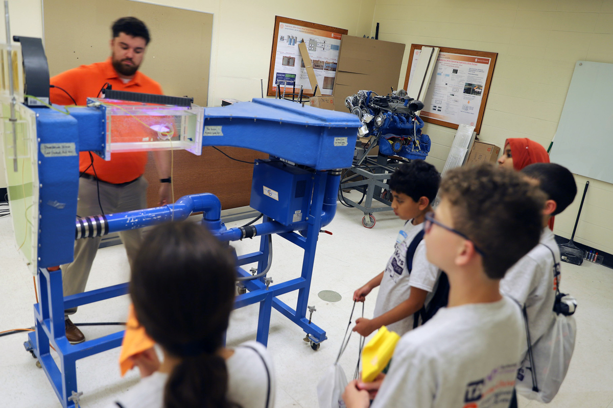 A group of young students observes a large blue mechanical testing apparatus or wind tunnel device in what appears to be an engineering laboratory or classroom. An instructor in an orange shirt operates the equipment in the background. The blue machine has various tubes, chambers, and mechanical components mounted on a wheeled frame. Several students wearing white shirts are gathered around watching the demonstration, with one student holding what appears to be a yellow object. Educational posters and materials are visible on the walls behind them.