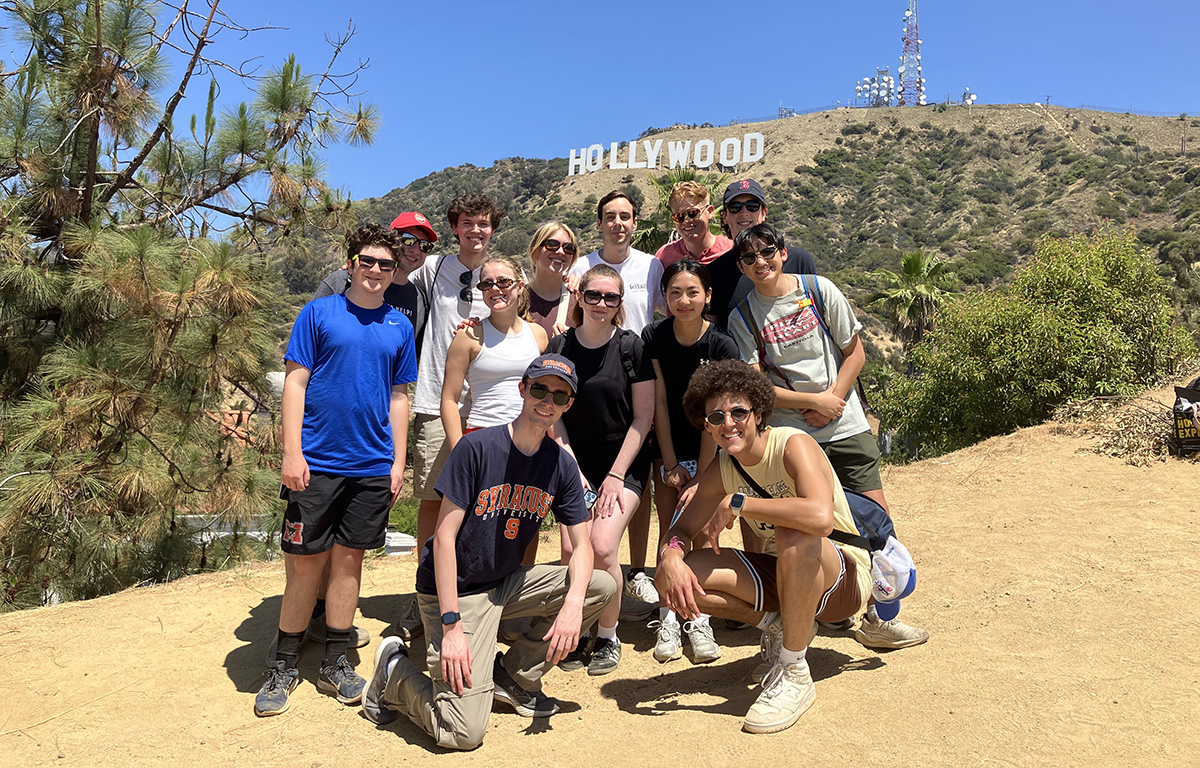 Group of 13 people posing on a dirt path with the Hollywood sign in the background.