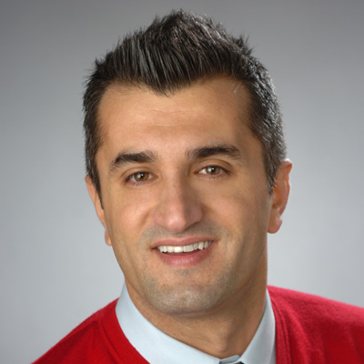 A person with short, dark hair wearing a red sweater over a light blue collared shirt against a light blue background.