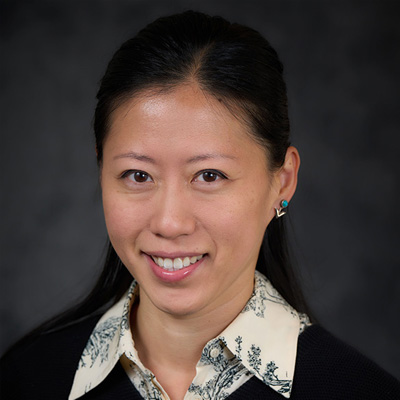 A person with long dark hair wearing a black top over a white collared shirt with a floral pattern against a dark background.
