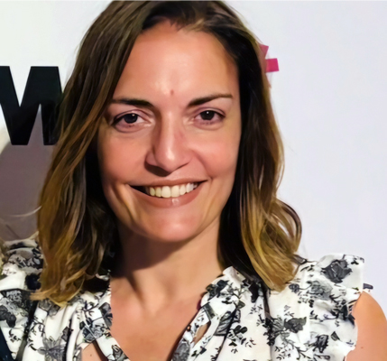 A person with shoulder-length brown hair with blonde highlights smiles brightly at the camera. They are wearing a white top or dress with a black floral pattern. The photo appears to be taken at an event, with a white backdrop visible behind her that shows partial text or logos.