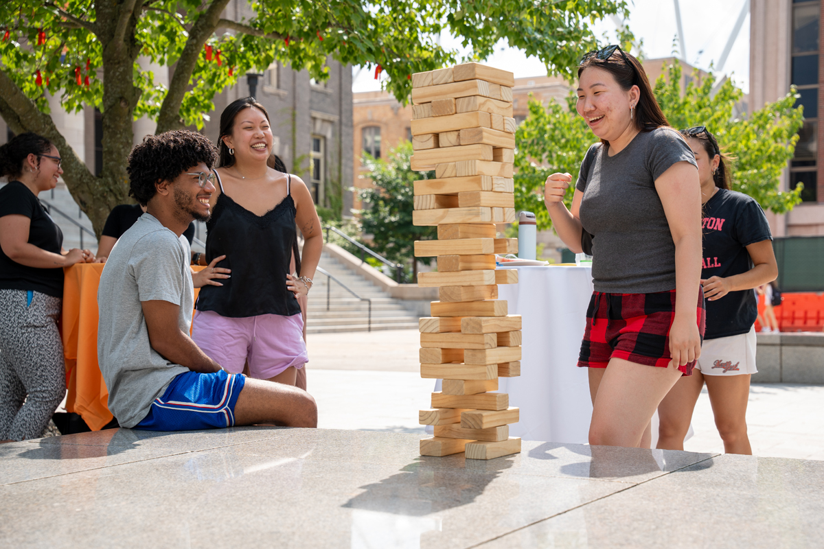 Group of people gathered outdoors around a large wooden Jenga tower in a public space with trees and buildings in the background. One person is interacting with the tower while others stand or sit nearby, engaged in a social and leisurely activity.