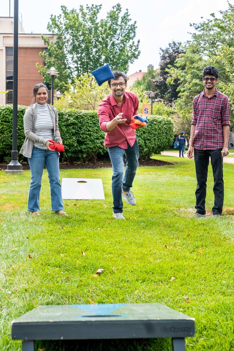 Three people play a game of cornhole on the Quad. One person is mid-throw with a blue bean bag aimed at the cornhole board, while the other two hold red and blue bean bags. The background features green bushes, trees, and part of a building.
