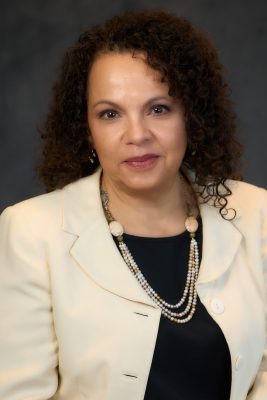 A person with curly hair wearing a light-colored blazer and a necklace, standing against a dark background.