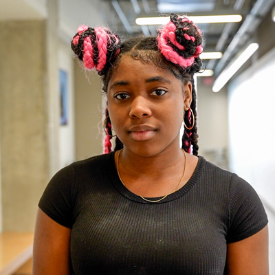 Person with pink and black braided hair in two buns, wearing a black ribbed shirt and necklace, standing in an industrial-style hallway with ceiling lights and blurred background.