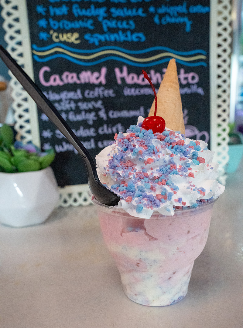 Colorful ice cream dessert in a clear plastic cup with layers of pink and white ice cream, topped with whipped cream, blue and pink sprinkles, a cherry, and an upside-down cone; black spoon inserted; background features a chalkboard menu with handwritten dessert items.