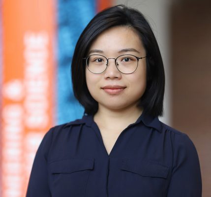 A young person with shoulder-length black hair and round, thin-framed glasses smiles gently at the camera. They are wearing a dark navy blue button-up shirt or blouse with chest pockets. The background shows the same modern interior space with blurred orange and blue architectural elements, suggesting a contemporary office or building environment.