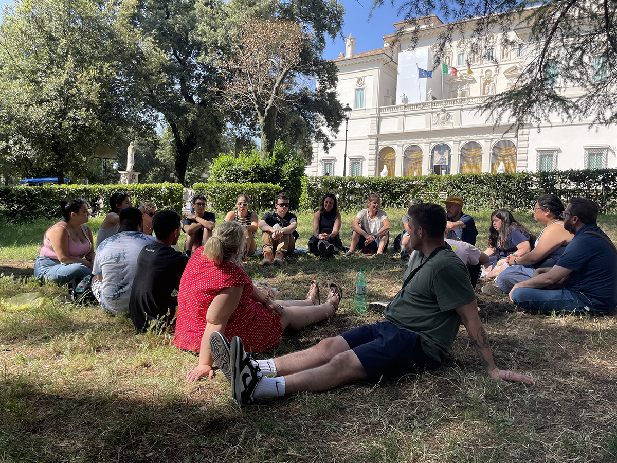A group of people sits in a circle on the grass in an outdoor area. Behind them is a large building with multiple windows and several flags displayed on its facade. Trees surround the area, and the group is engaged in discussion. 