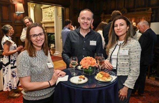 Three people attending an after-discussion reception pose and smile facing the camera among others around them