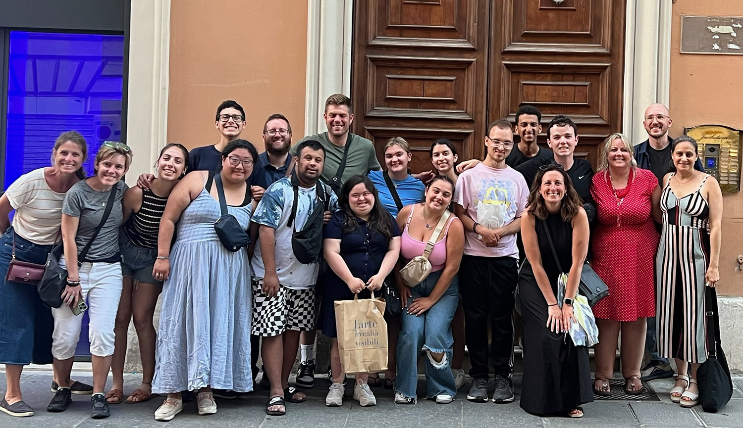 A large group of people poses in front of a large wooden door. Some are kneeling or squatting in the front row, while others stand behind them. They are dressed casually, and one person holds a shopping bag with the text 'l'arte rende visibili.' The background features an orange wall and a blue-lit window on the left.