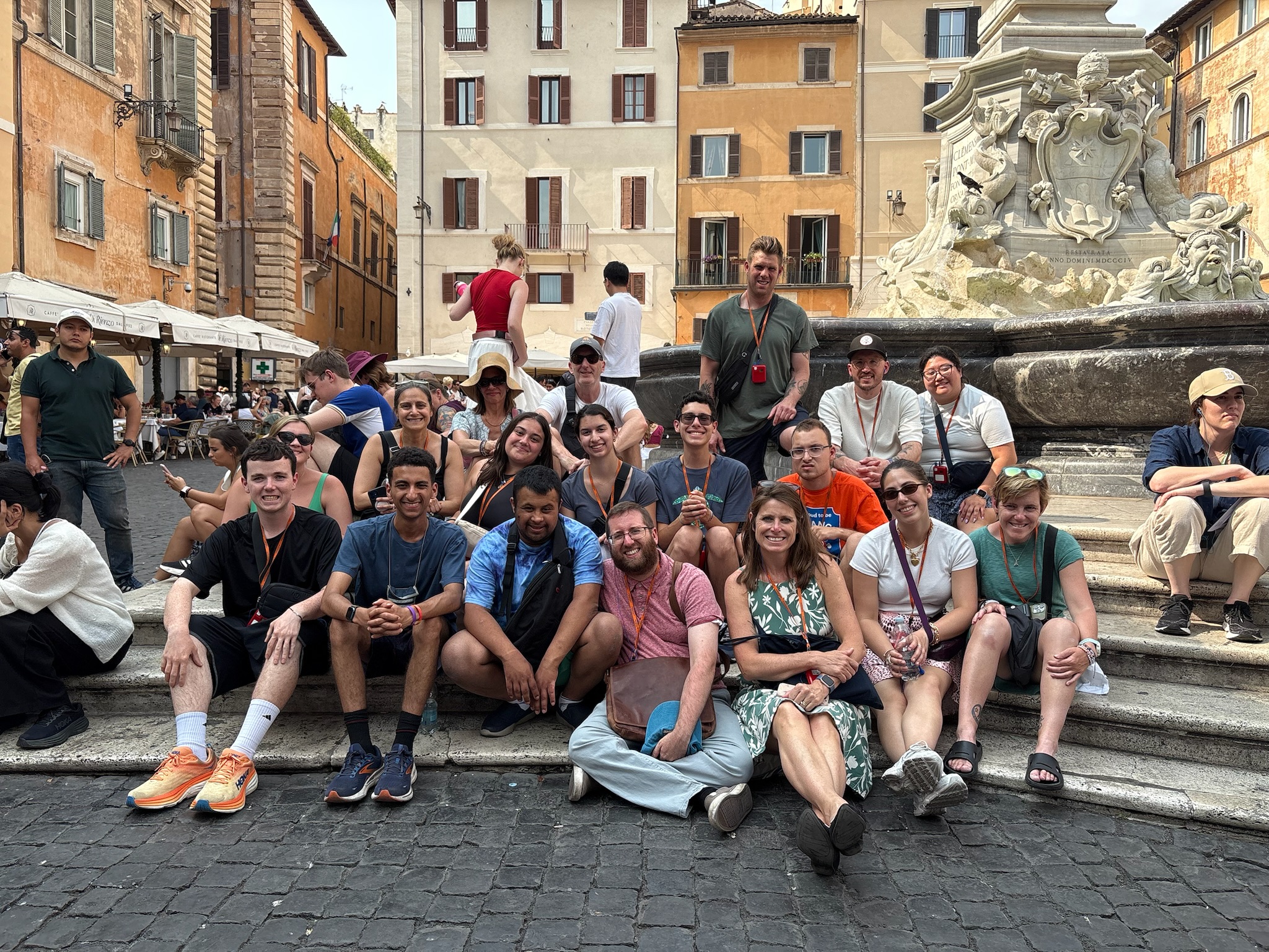 A group of people sits on the steps of an ornate fountain in an outdoor plaza. The background features multi-story buildings with balconies and windows. Other people are walking or standing around the plaza, and tables with umbrellas are visible on the left. The ground is paved with cobblestones.