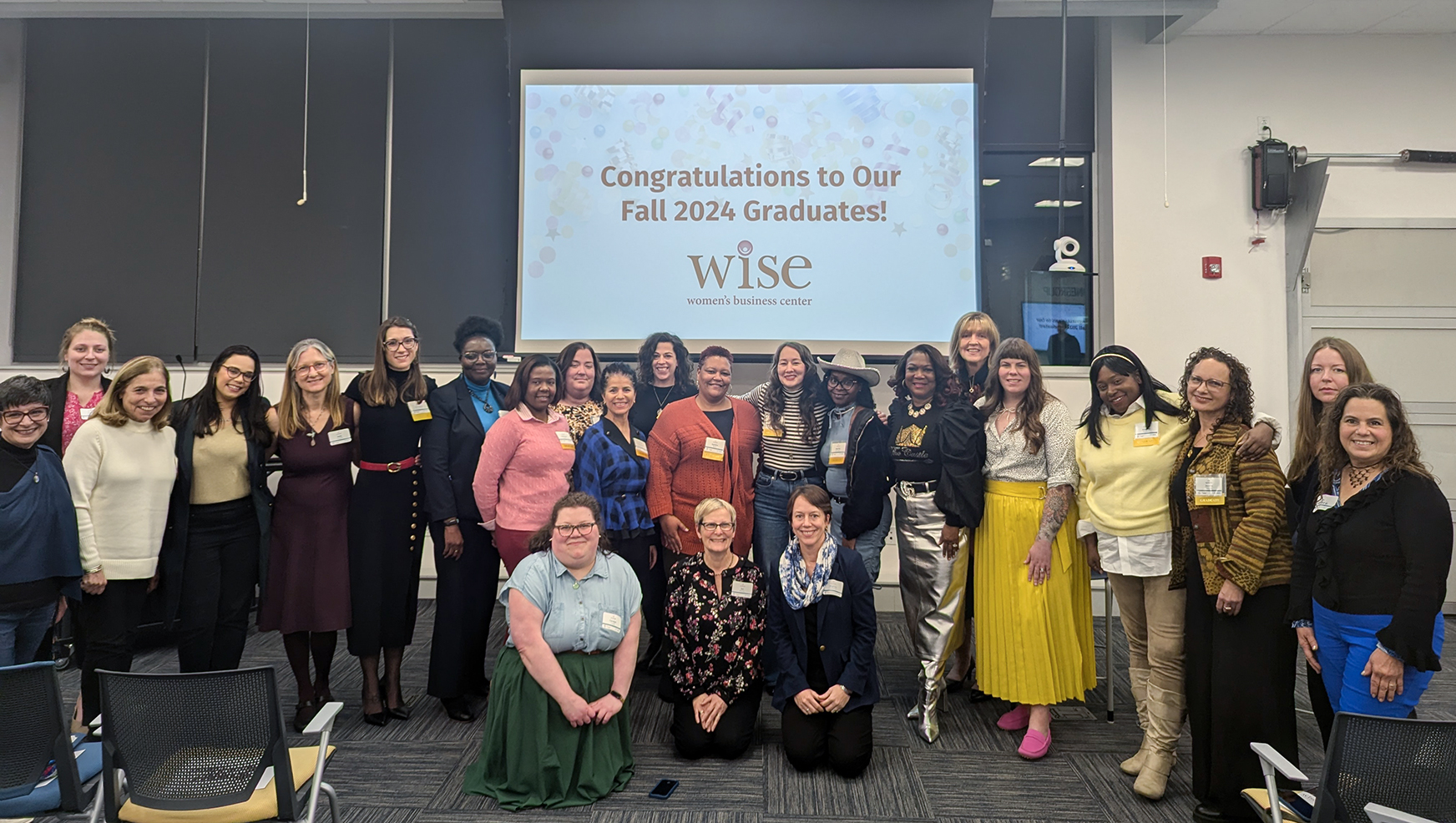 group of people posing in a classroom in front of a screen that states Congratulations to Our Fall 2024 Graduates, WiSE