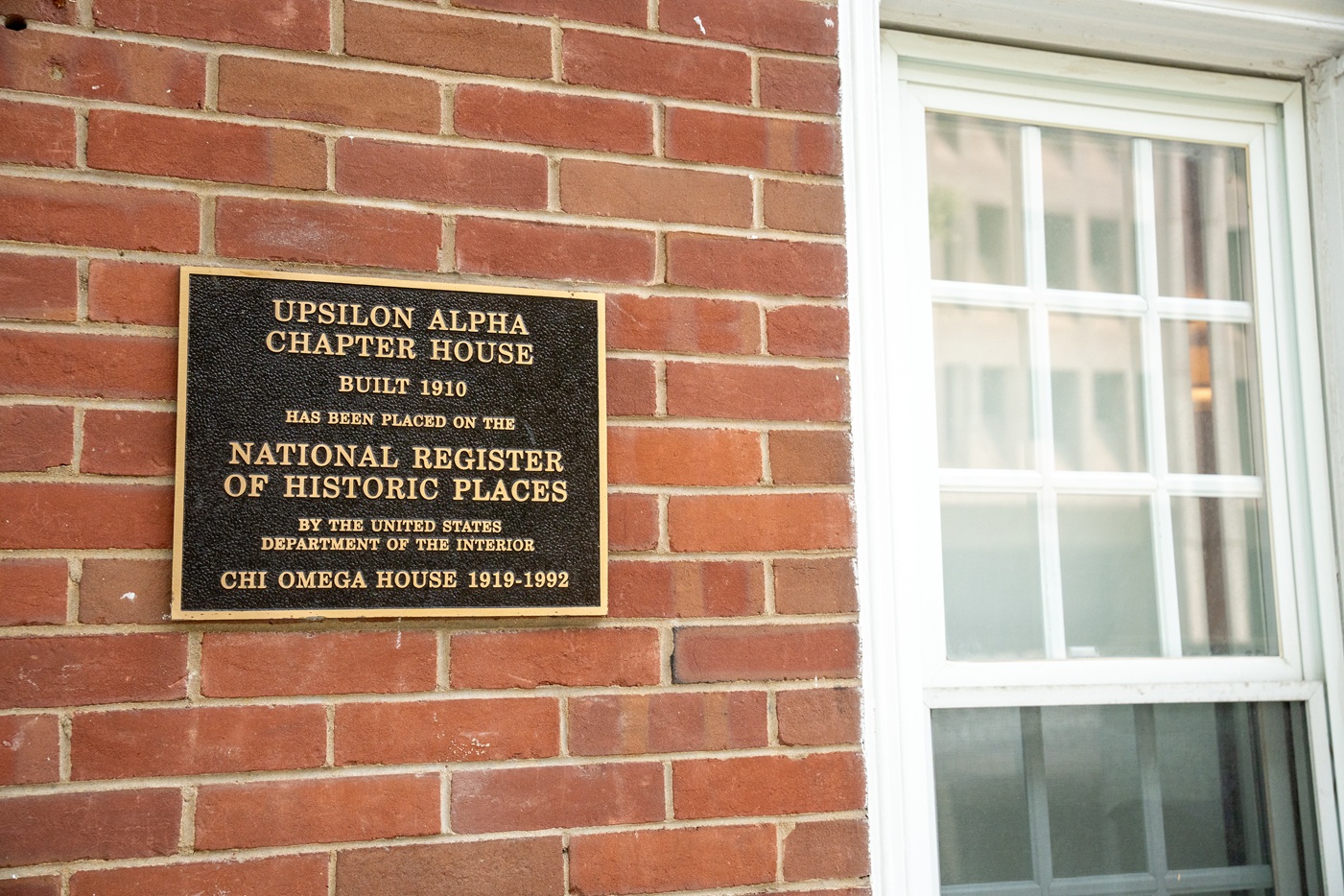 A plaque mounted on a brick wall next to a window. The plaque reads: "UPSILON ALPHA CHAPTER HOUSE BUILT 1910 HAS BEEN PLACED ON THE NATIONAL REGISTER OF HISTORIC PLACES BY THE UNITED STATES DEPARTMENT OF THE INTERIOR CHI OMEGA HOUSE 1919–1992."