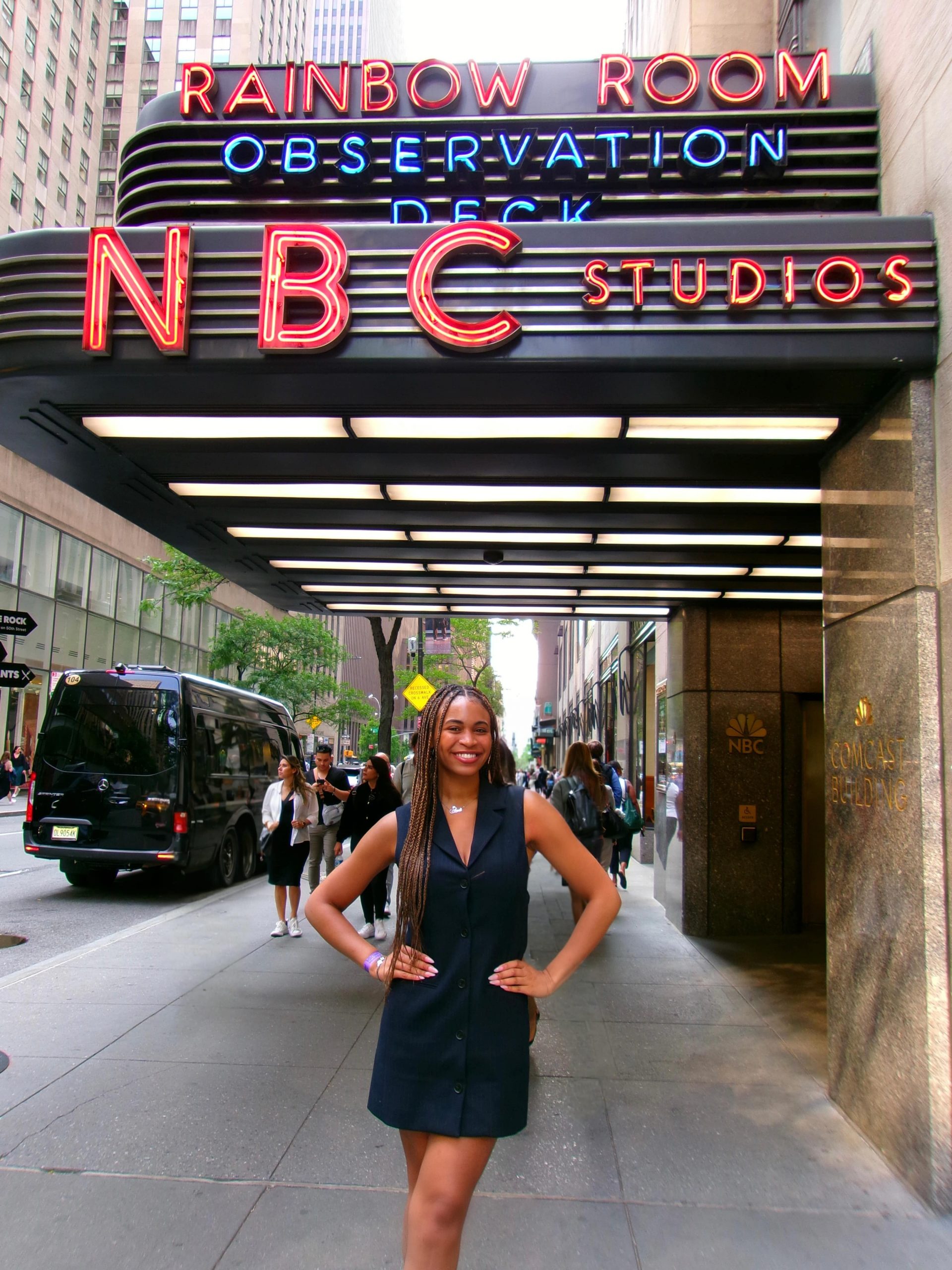 A student wearing a navy blue dress poses with hands on her hips in front of the NBC Studios entrance in New York City, with the iconic "Rainbow Room Observation Deck NBC Studios" neon sign visible above.