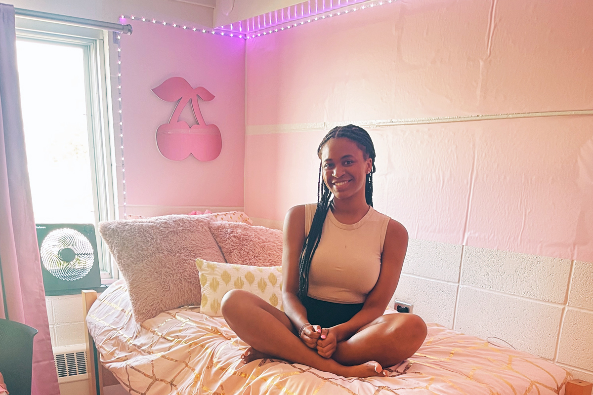 A student sits cross-legged on a bed with golden bedding in a pink-themed bedroom, smiling at the camera. The room features pink walls with cherry decorations and a small fan by the window.