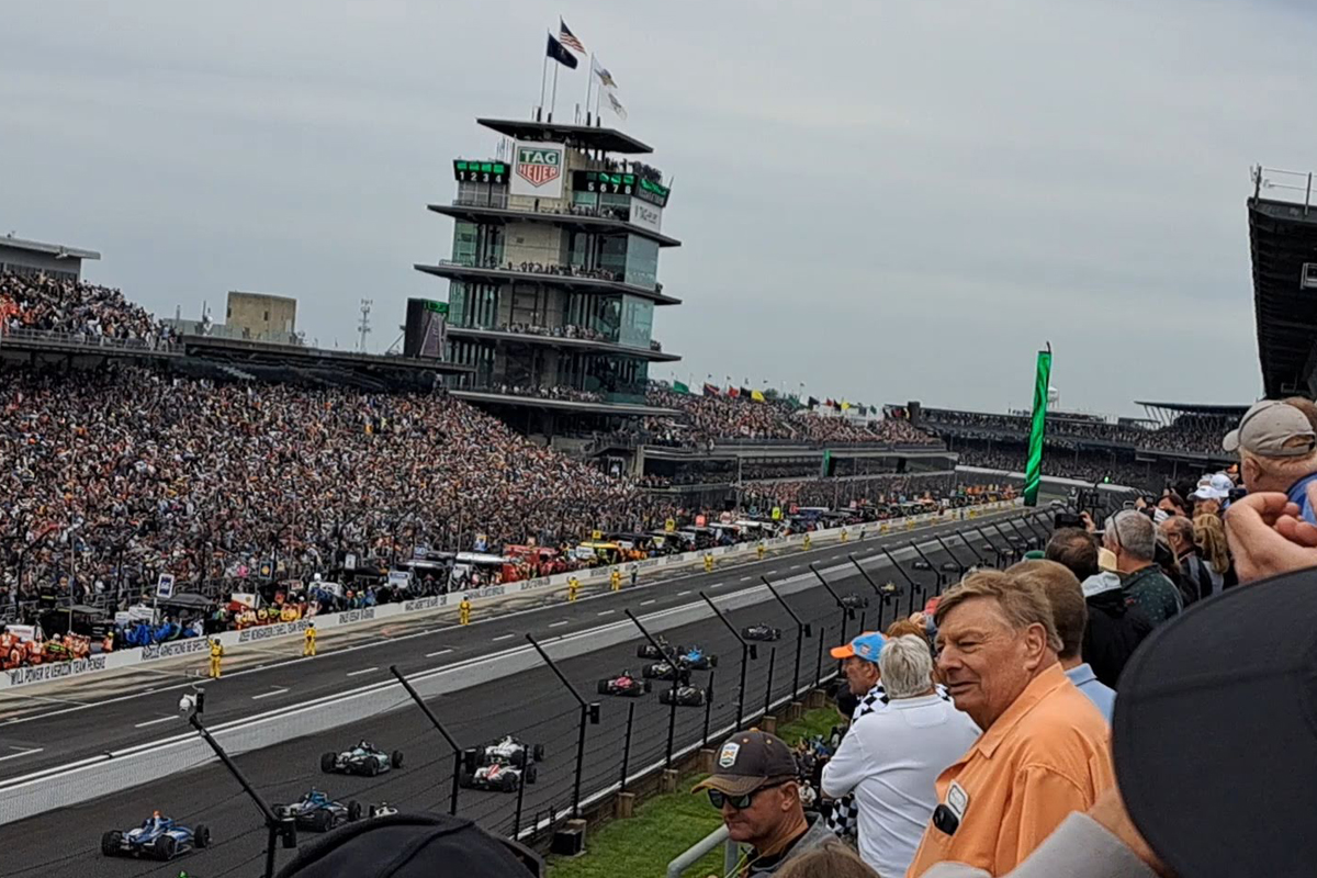 Indianapolis Motor Speedway during race with packed grandstands, iconic Pagoda control tower visible, and IndyCars racing on the famous oval track.