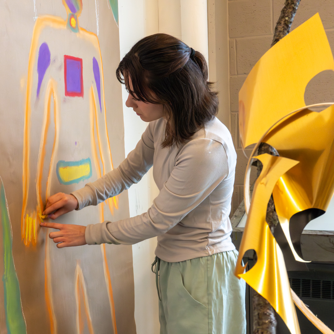 Person drawing an abstract human figure with colorful shapes on a large paper, using yellow chalk near a yellow sculpture.