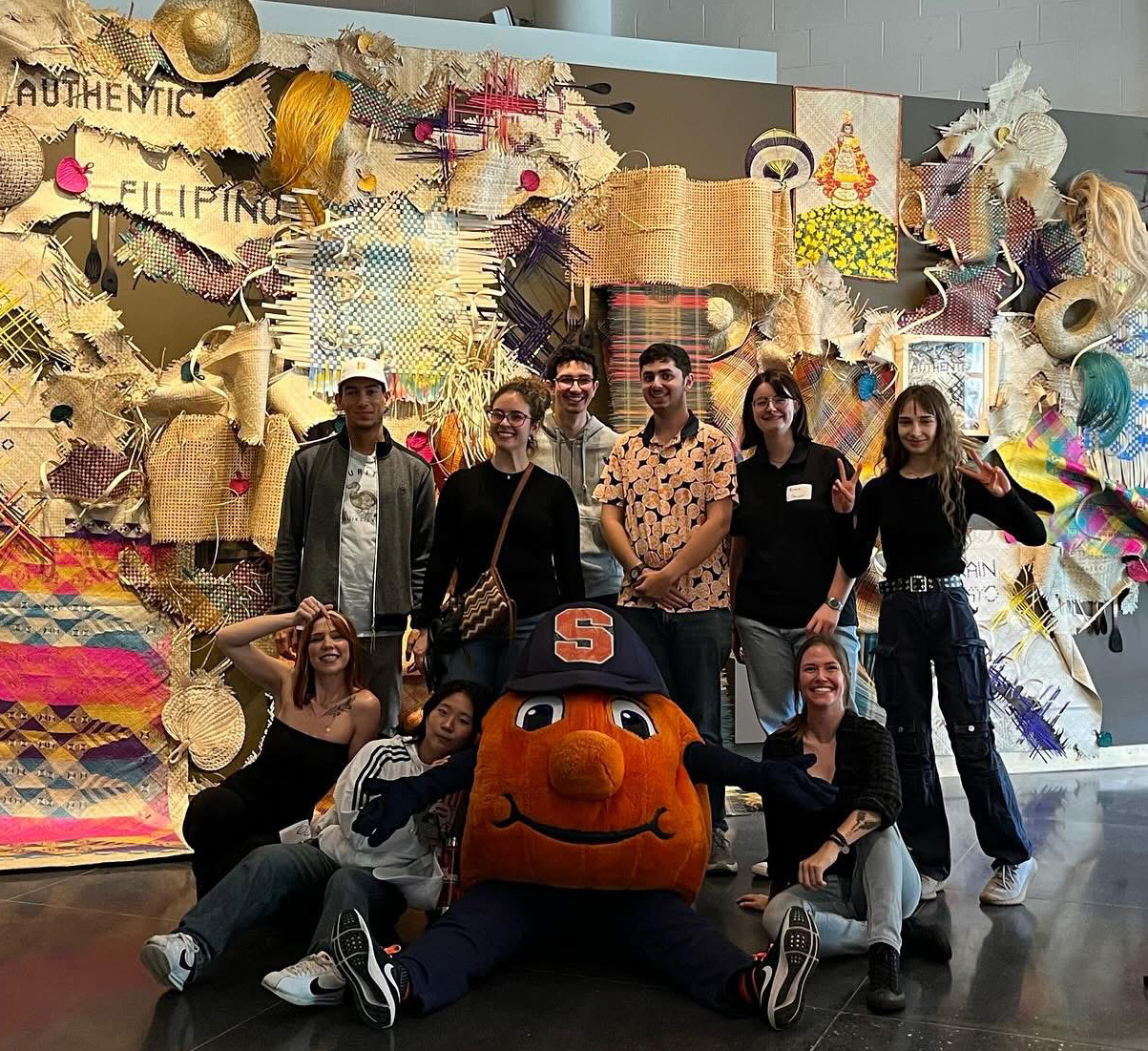 Group of nine people pose with Otto the Orange mascot in front of a vibrant art installation labeled 'Authentic Filipino' with woven textiles and decorations.