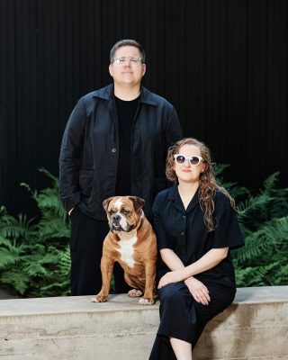 Professional portrait of two people in matching navy blue outfits with their brown and white bulldog, posed against a black backdrop with green foliage accents.