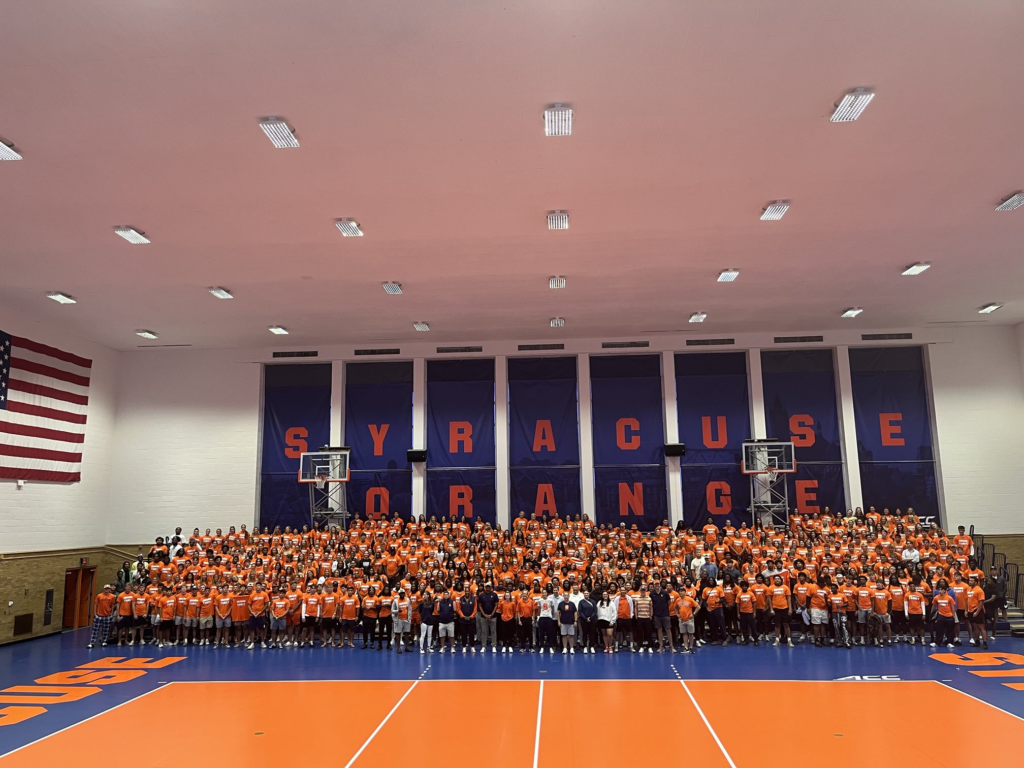 Large group in orange shirts poses for a photo on a Syracuse University basketball court, with "SYRACUSE ORANGE" banners, hoops, and ACC branding visible in a brightly colored gymnasium.