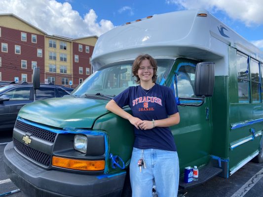 Young woman in Syracuse t-shirt and jeans stands alongside a green and white bus