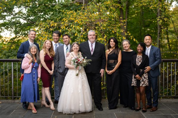 Wedding photo, a bride and groom in the center with nicely dressed guests flanking them. Fall foliage appears in the background