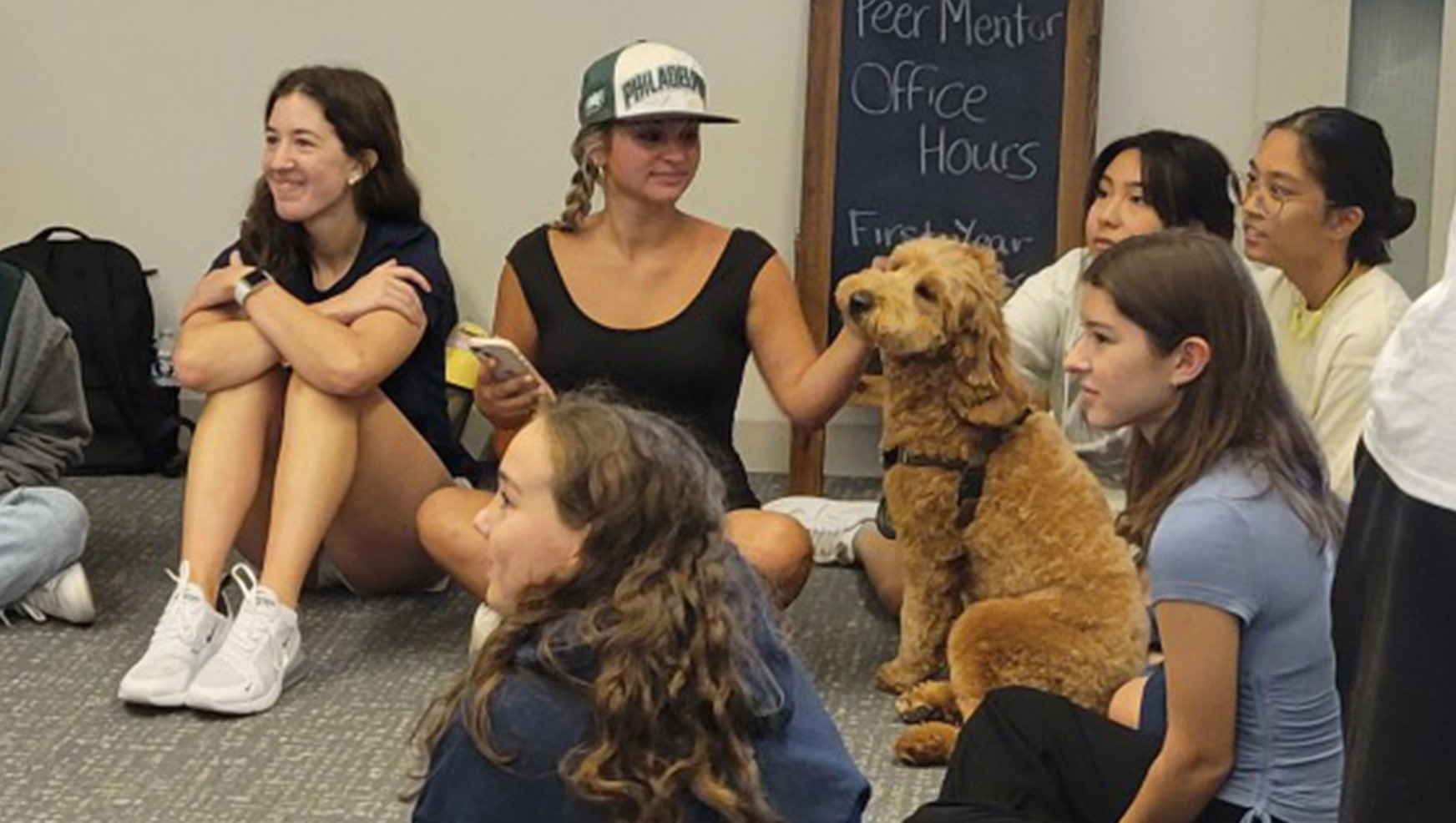 A group of students sits on the floor surrounding a brown dog.