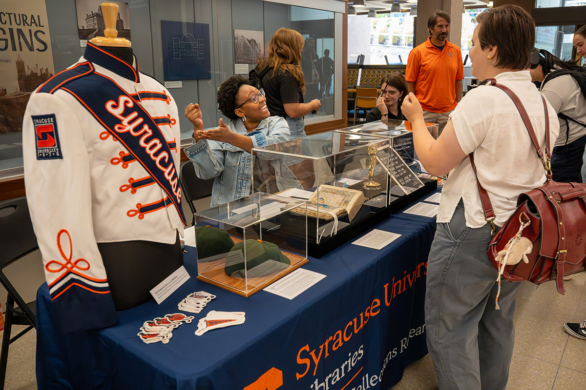 Display table with Syracuse University marching band uniform, historical artifacts, and informational materials, surrounded by attendees at an event.