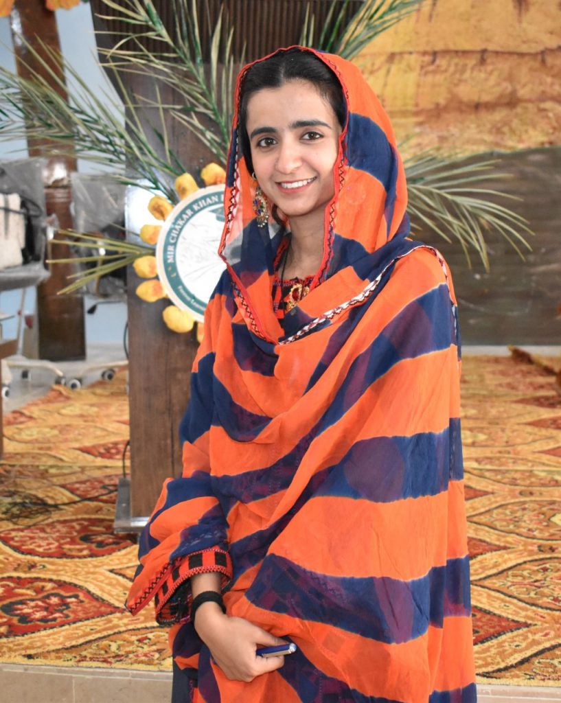 A young person wearing a colorful orange and navy blue headscarf smiles at the camera, with palm fronds and decorative elements visible in the background.