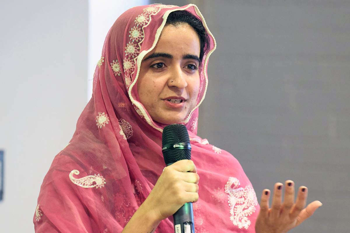 A young person wearing an ornate pink headscarf with gold embroidery speaks into a microphone while gesturing with her hand.