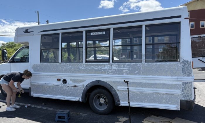 Person working near the rear wheel of a partially stripped white minibus with large windows. Tools, wood pieces, and a blue step stool are scattered on the ground nearby."
