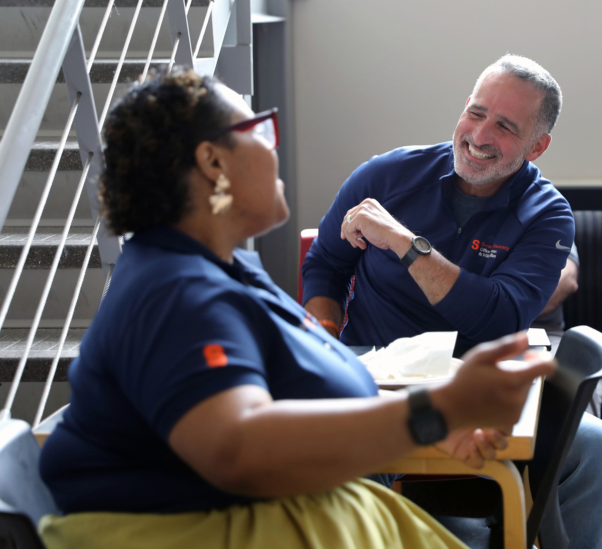 Two people in dark blue shirts with orange logos sit at a table conversing in a casual indoor setting with a staircase in the background.