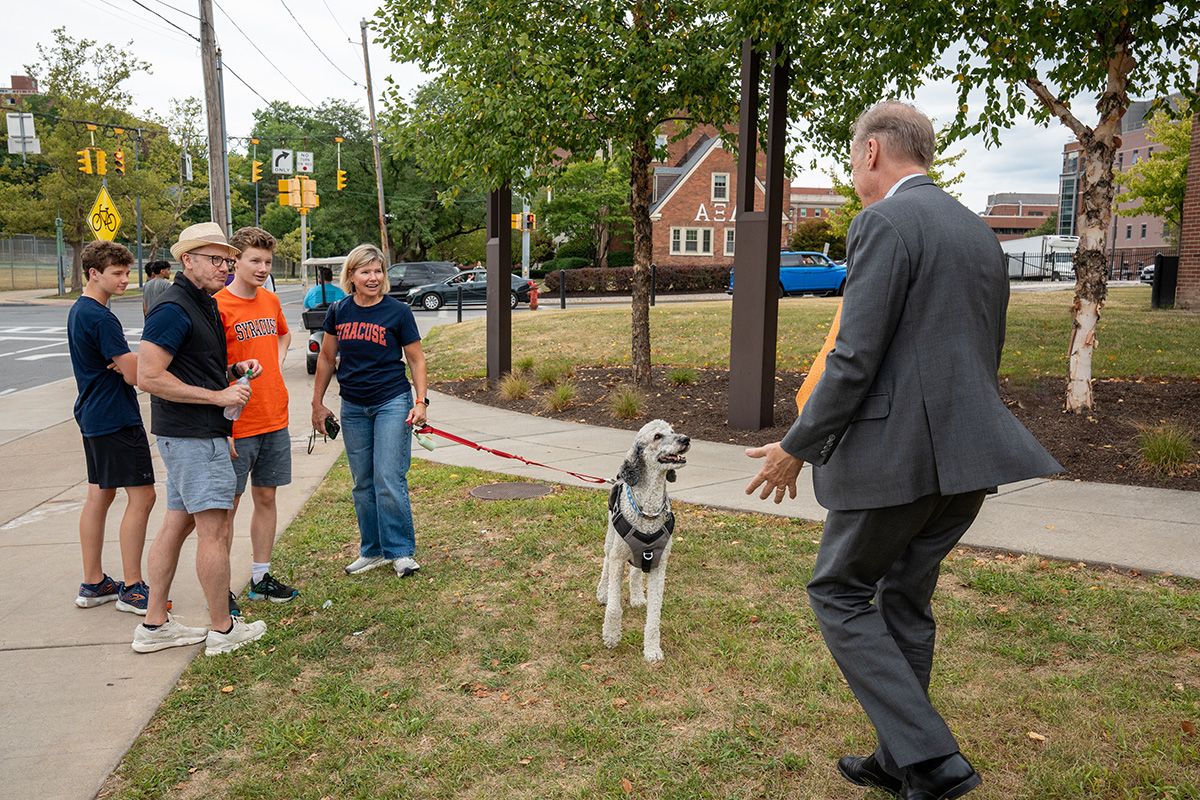 Group of people, some in Syracuse gear, standing on grass near a sidewalk with a dog on a leash and Chancellor Kent Syverud approaching.