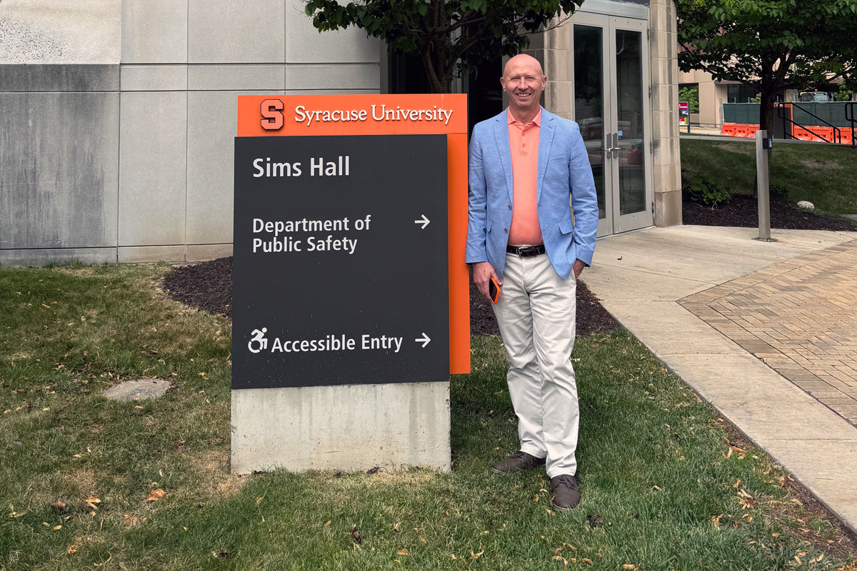 A person in a light blue blazer and orange shirt stands next to a Syracuse University directional sign for Sims Hall and the Department of Public Safety on campus.