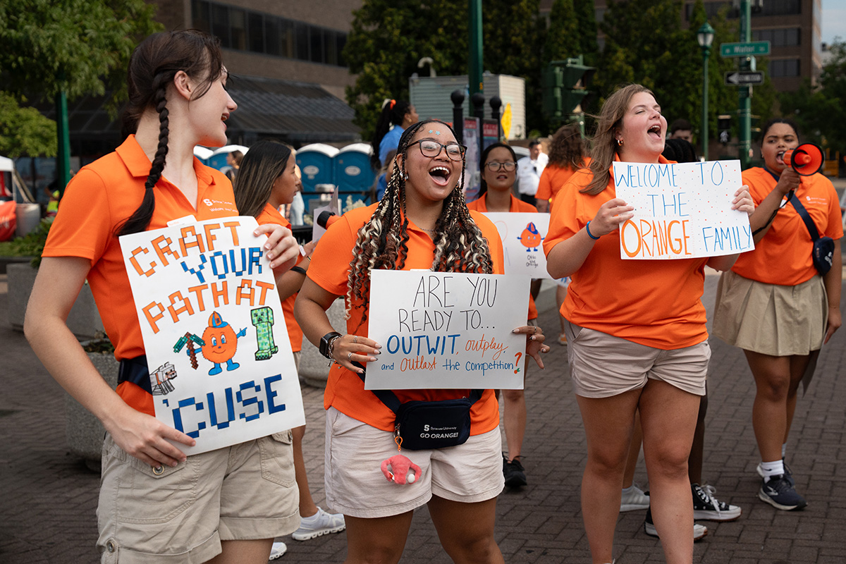 Group of people in orange shirts holding welcoming signs with Syracuse-themed messages, standing outdoors near trees and buildings.