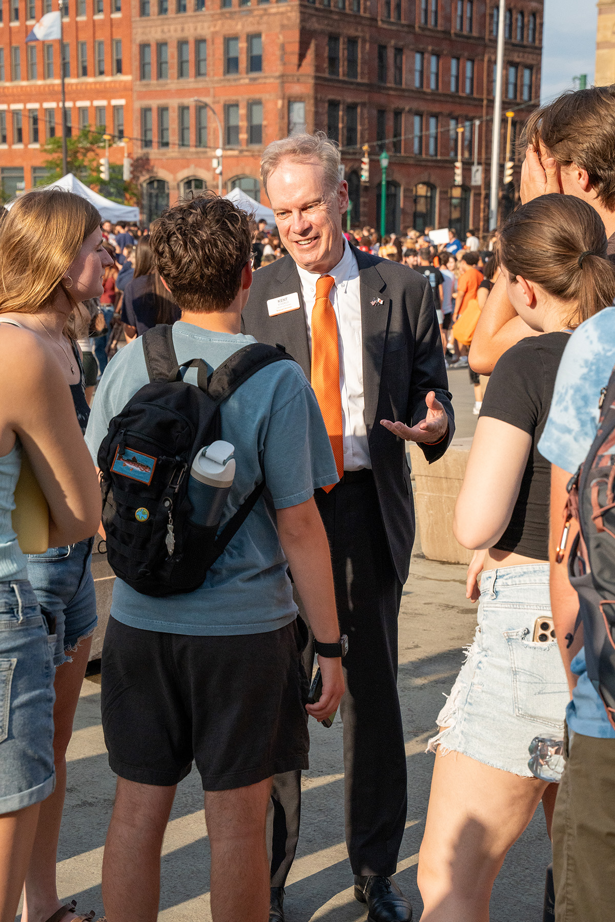 Chancellor Kent Syverud in a suit with an orange tie and name tag speaks to a group outdoors in a city setting, with brick buildings and a crowd in the background.