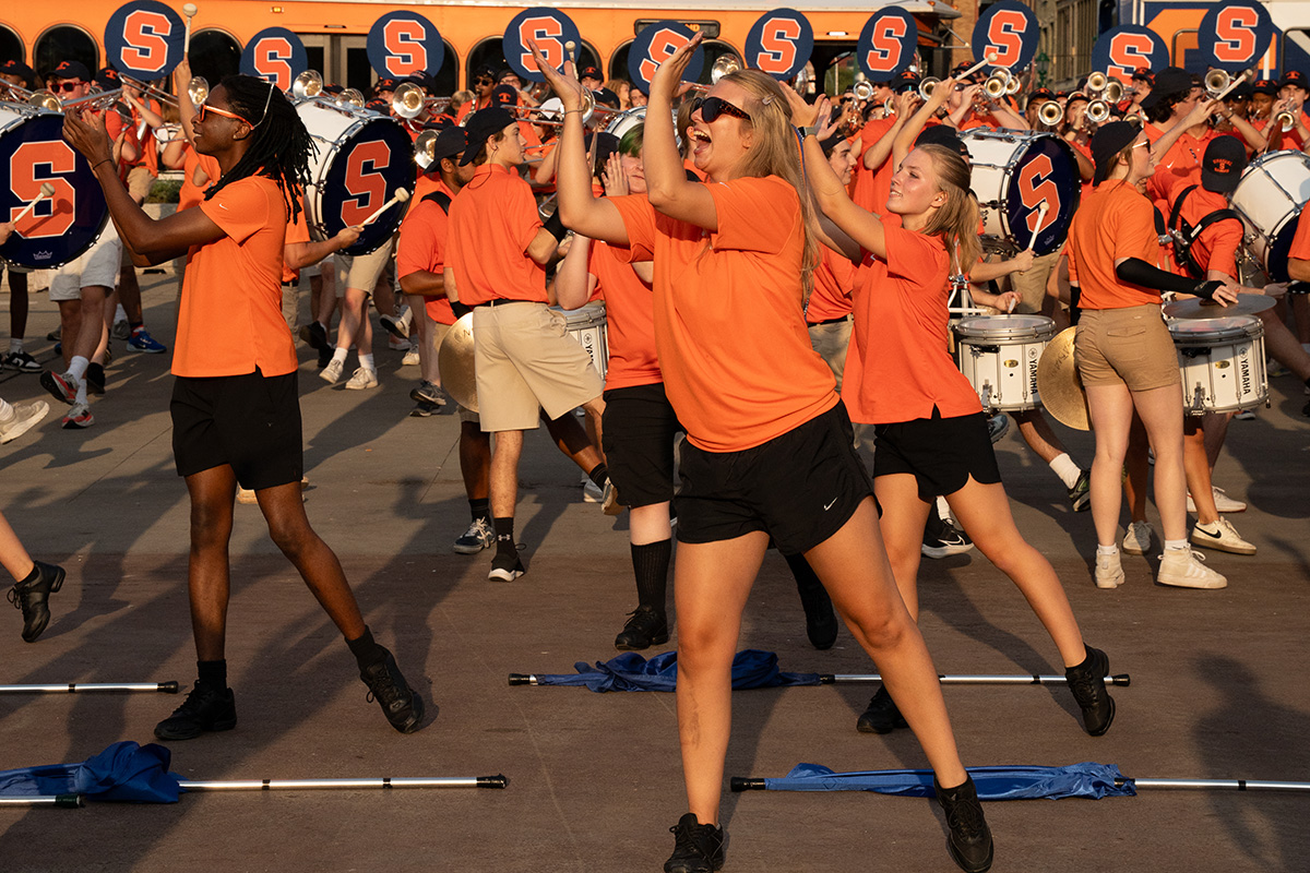 Group in orange shirts performing a dance routine outdoors, with a marching band in blue uniforms and orange 'S' letters in the background.