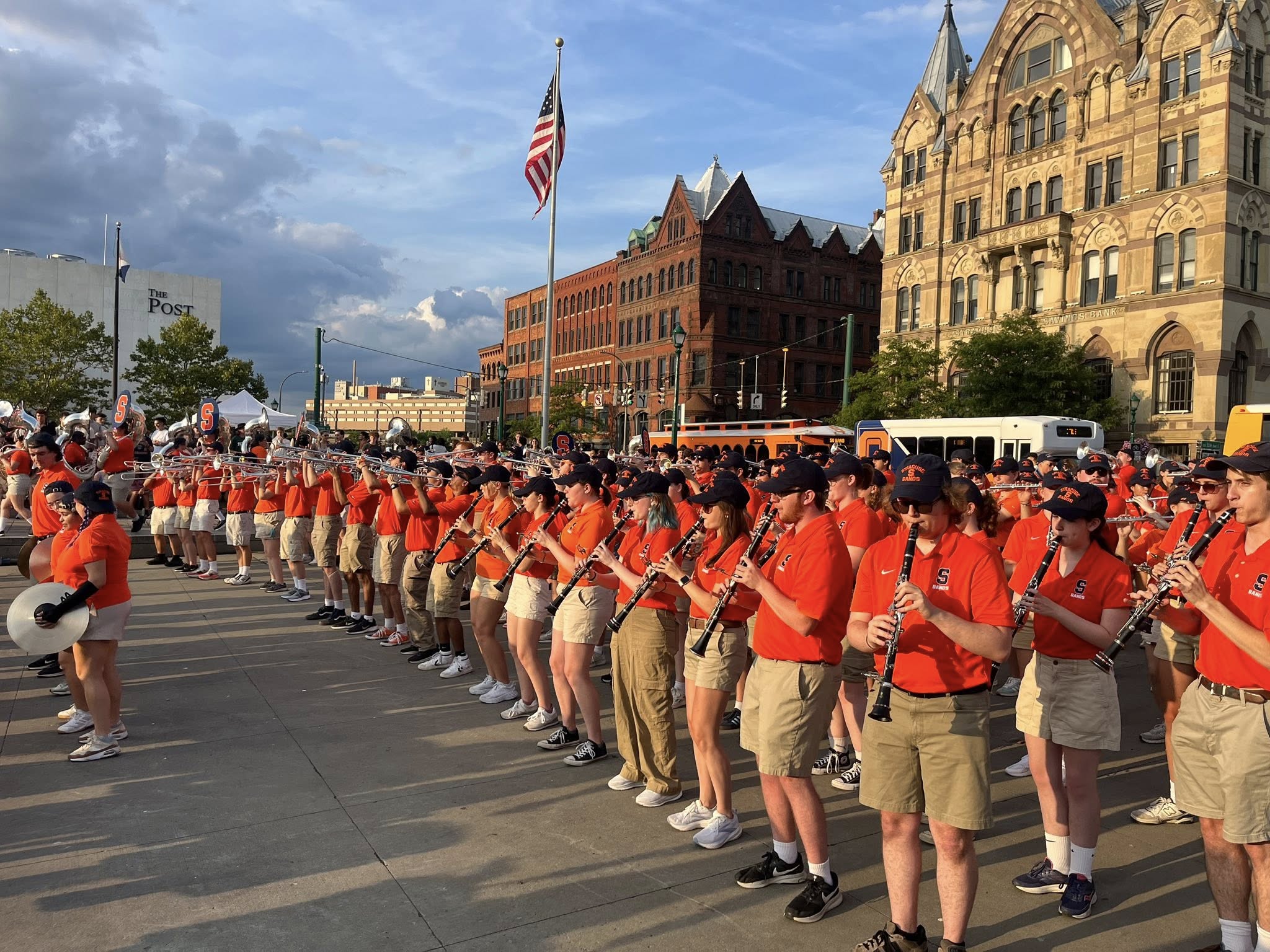 Marching band in orange shirts and navy caps performs with snare drums and blue flags in a lively public plaza, surrounded by spectators, a tall monument, and city buildings.