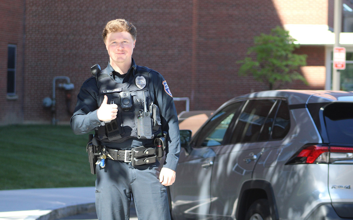 Campus safety officer giving a thumbs-up outdoors near a brick building and parked SUV, with greenery in the background.