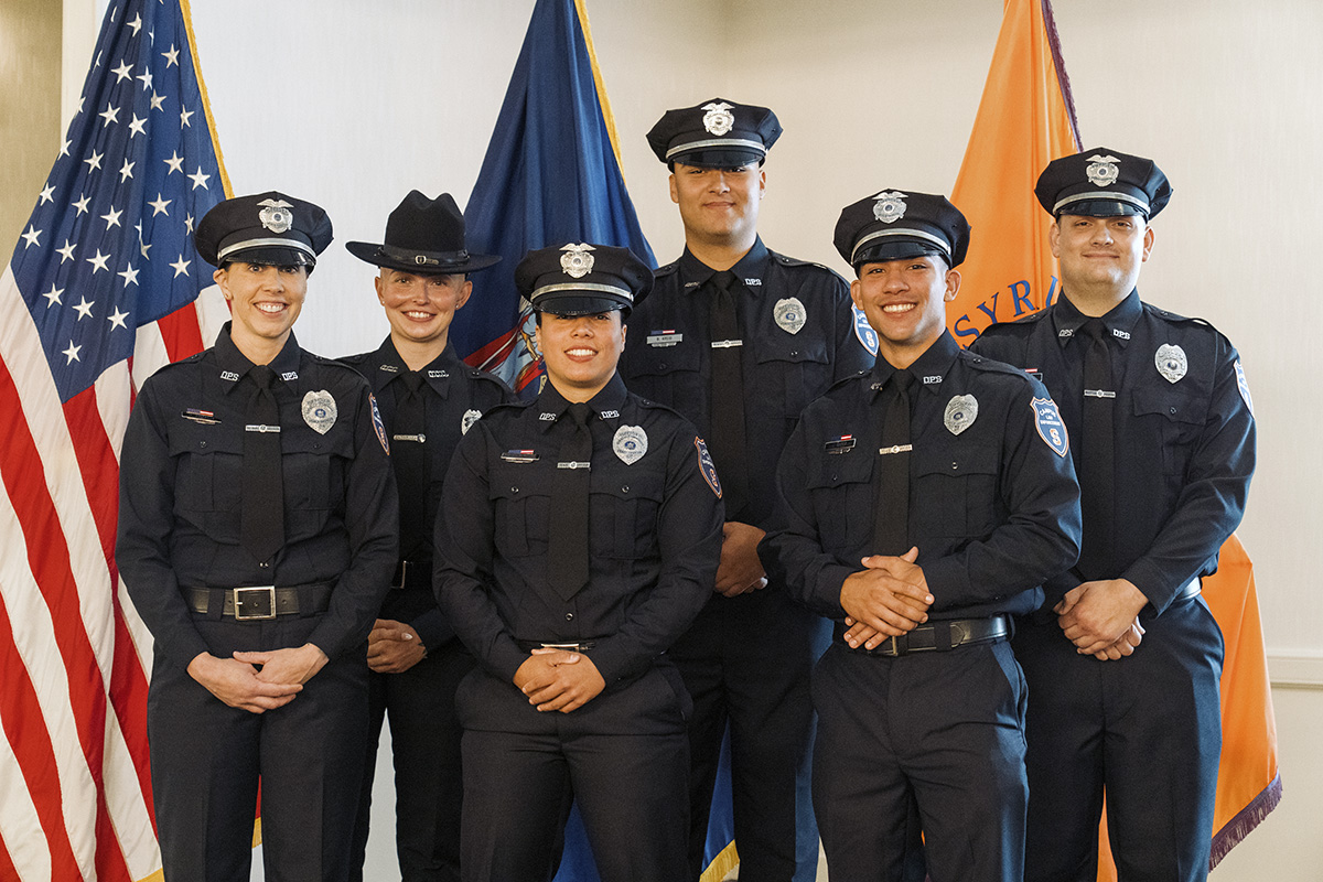 Six uniformed police officers standing in two rows in front of American, blue, and orange flags