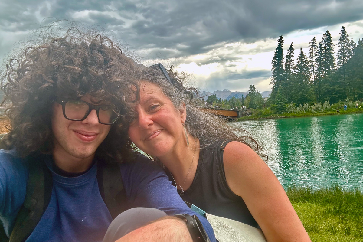 Two people with curly hair taking a selfie by a turquoise lake, mountains and evergreen trees visible in background under dramatic cloudy sky.
