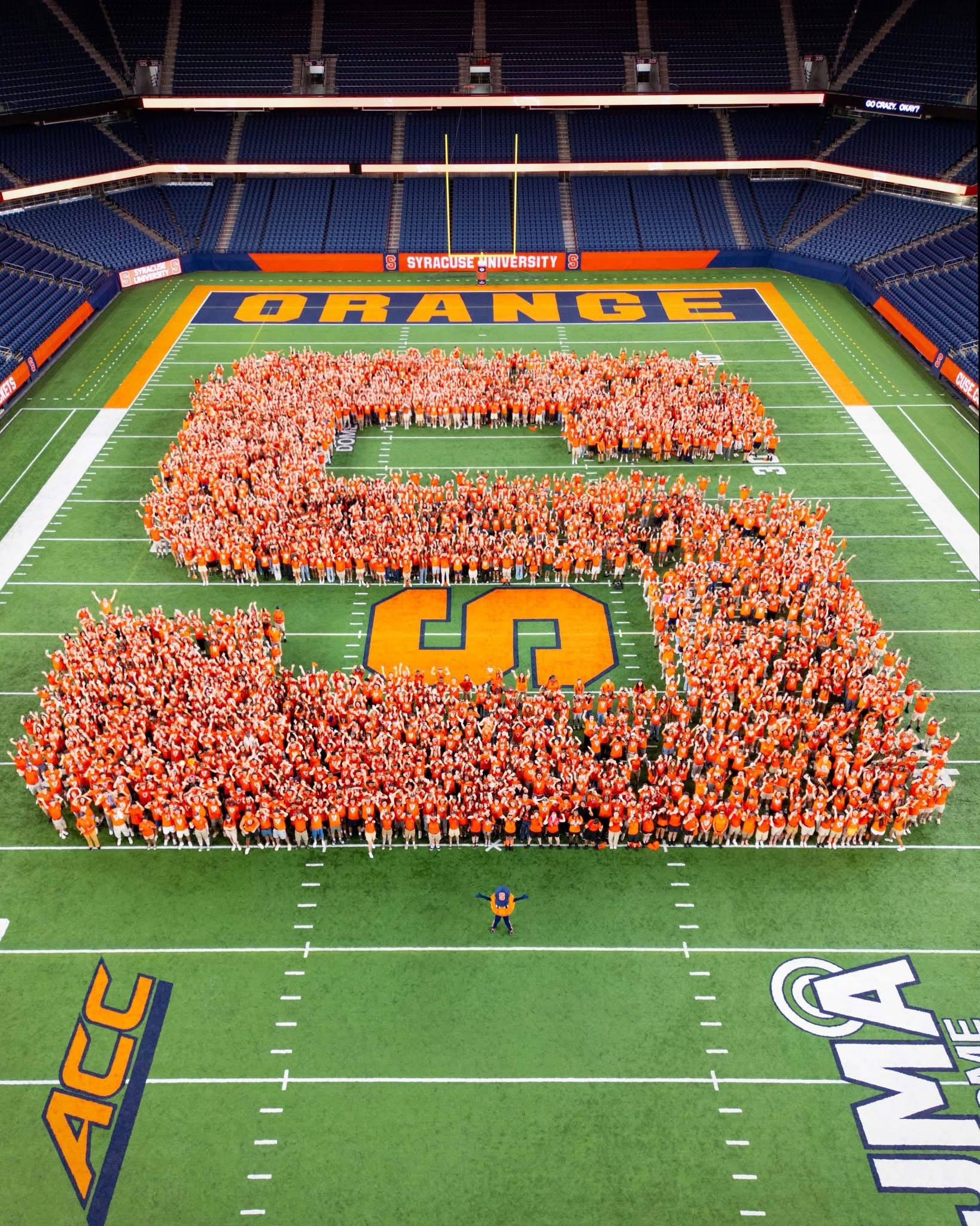 The members of the Class of 2029 in orange clothing form a large "S" on the football field at Syracuse University's JMA Wireless Dome.